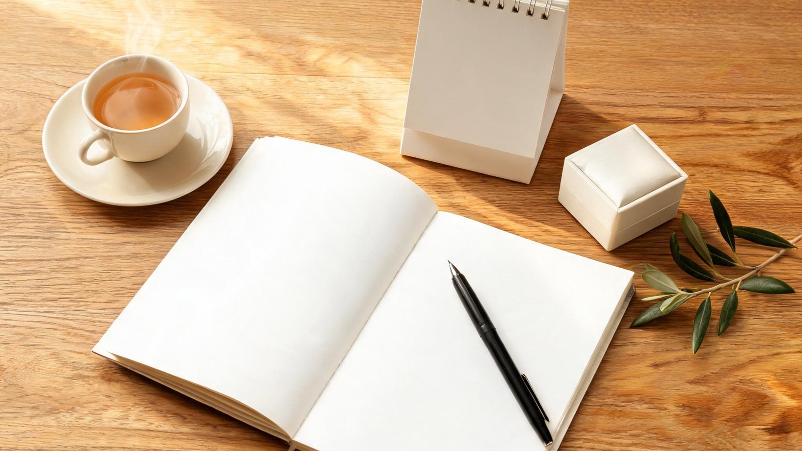 An open blank notebook, a pen, a blank desk calendar, a closed ring box, a cup of tea, and an olive sprig on a wooden table. Illustrative image, no text.